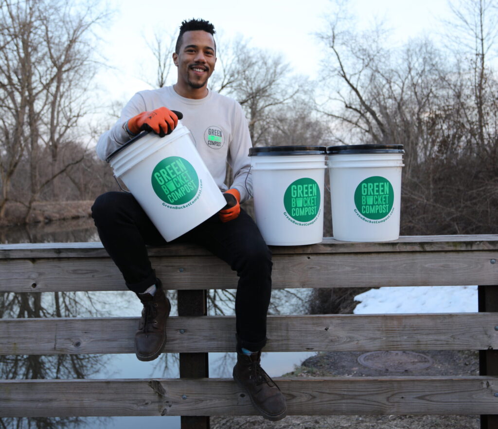 The founder of Green Bucket Compost, Isaiah, sitting on a bridge with three compost buckets